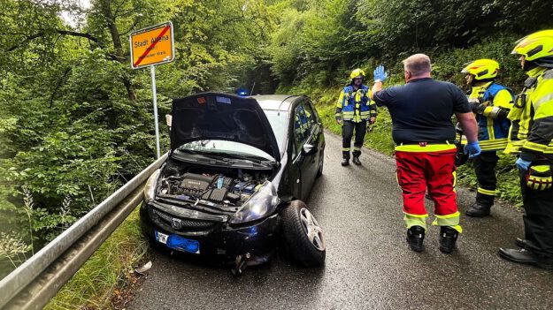 Foto: Carsten Menzel Ein schwarzes Auto steht mit geöffneter Motorhaube schräg an einer Leitplanke, das Vorderrad auf der Fahrerseite steht halb abgerissen von der Achse schräg aus dem Radkasten. Daneben stehen drei Feuerwehrleute und ein Mitarbeiter des Rettungsdienstes.