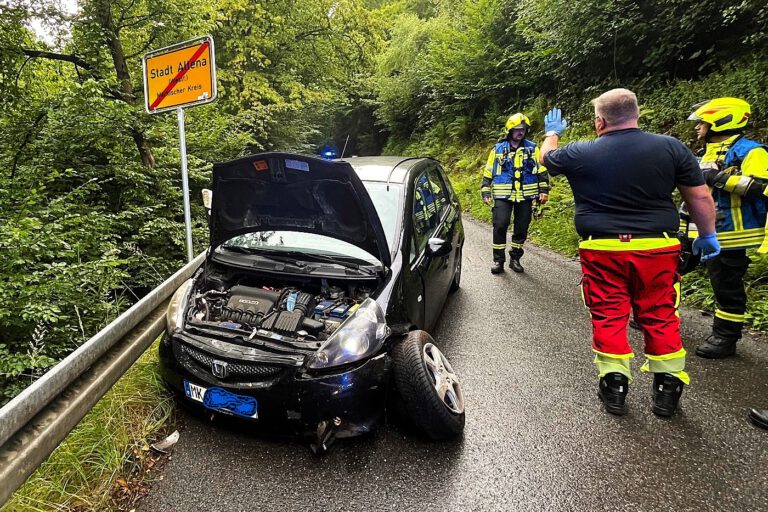 Ein schwarzes Auto steht mit geöffneter Motorhaube schräg an einer Leitplanke, das Vorderrad auf der Fahrerseite steht halb abgerissen von der Achse schräg aus dem Radkasten. Daneben stehen drei Feuerwehrleute und ein Mitarbeiter des Rettungsdienstes.