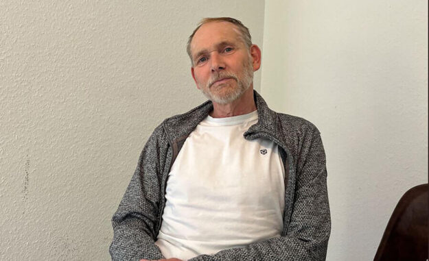 Foto: Kevin Kretzler Henrik Strübli sitzt in weißem T-Shirt und grauer Strickjacke vor einer weißen Wand.