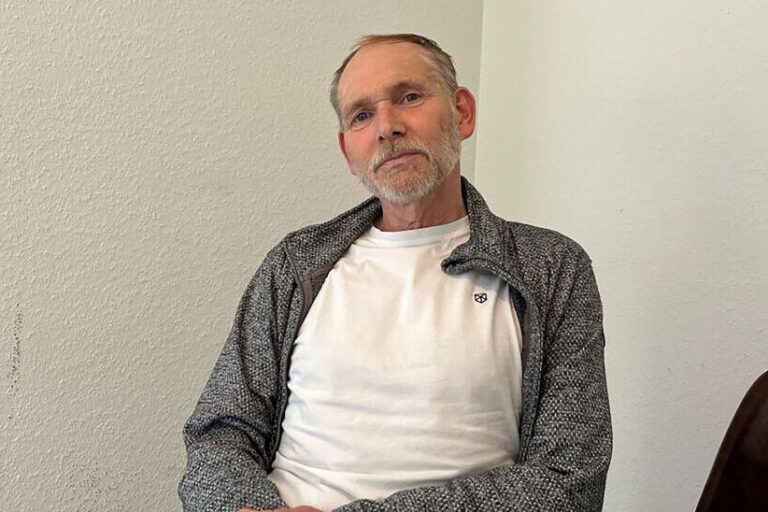 Henrik Strübli sitzt in weißem T-Shirt und grauer Strickjacke vor einer weißen Wand.