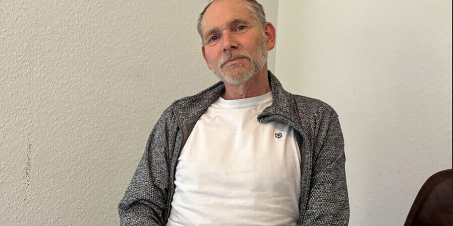 Henrik Strübli sitzt in weißem T-Shirt und grauer Strickjacke vor einer weißen Wand.