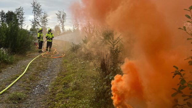Foto: Carsten Menzel Aus einem Waldstück steigt oranger Rauch auf. Zwei Feuerwehrleute löschen mit einem D-Rohr.
