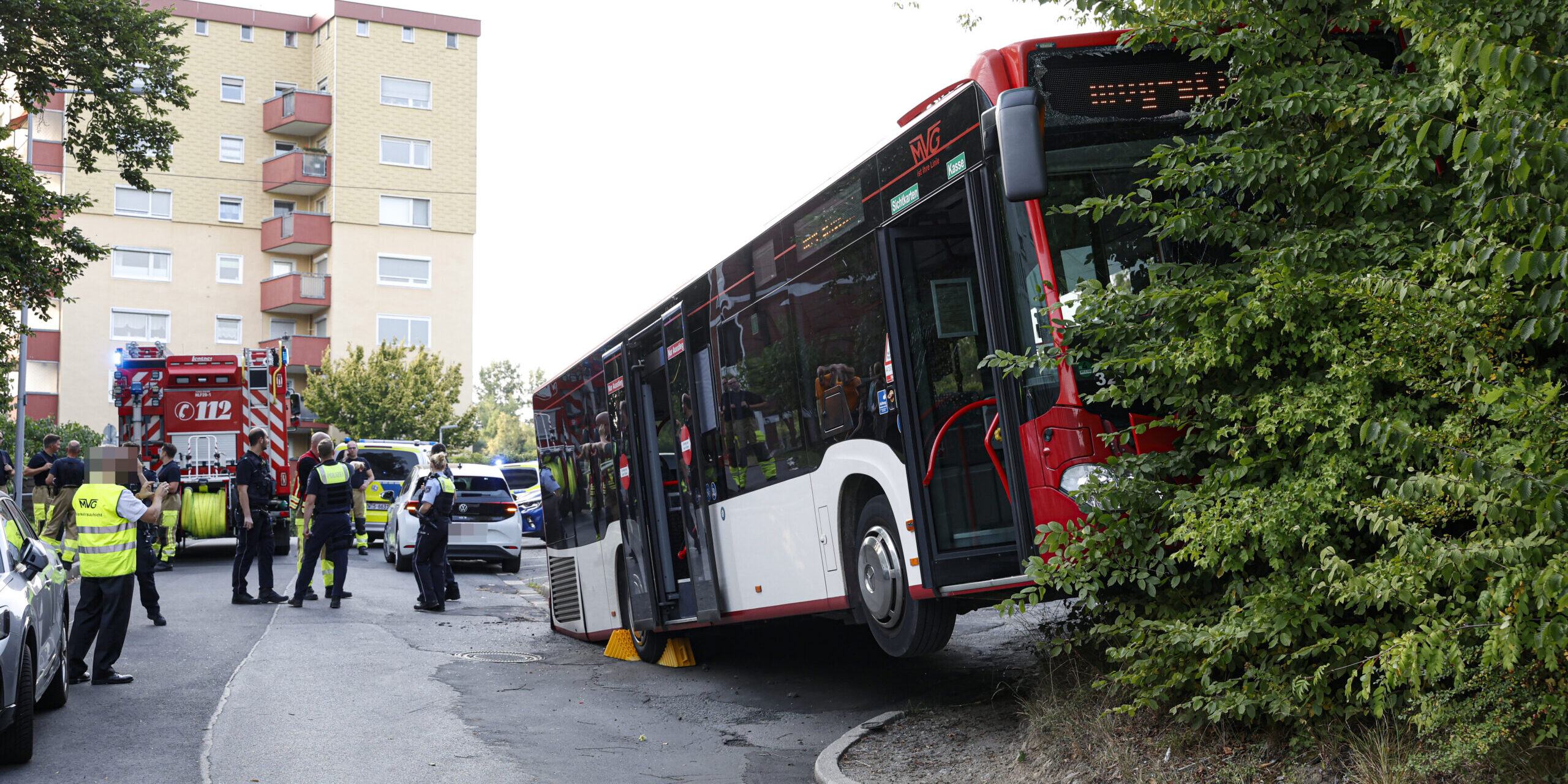 Ein rot-weißer Linienbus hängt in einem Hang fest; das Heck setzt auf der Straße auf. Links davon steht ein Feuerwehrauto und Feuerwehrmänner, Polizisten und Mitarbeiter der Busgesellschaft, die sich beraten.