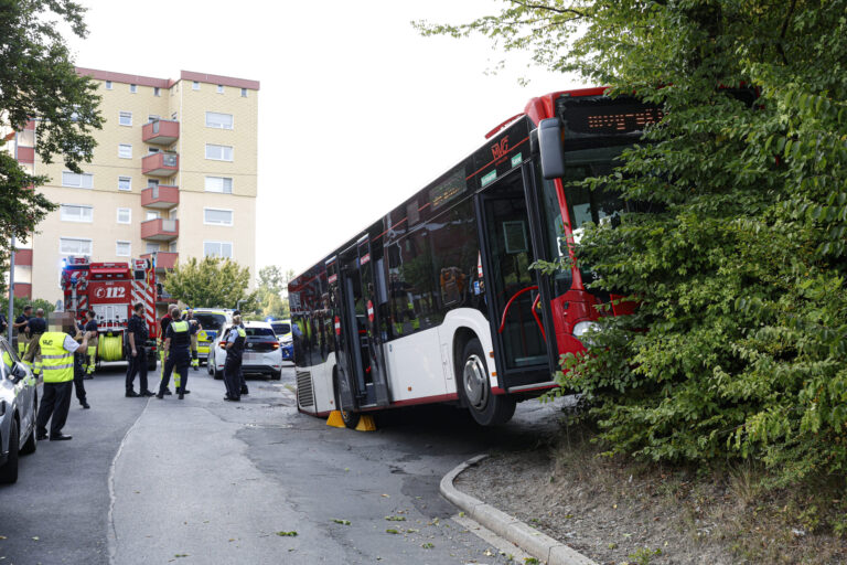 Ein rot-weißer Linienbus hängt in einem Hang fest; das Heck setzt auf der Straße auf. Links davon steht ein Feuerwehrauto und Feuerwehrmänner, Polizisten und Mitarbeiter der Busgesellschaft, die sich beraten.