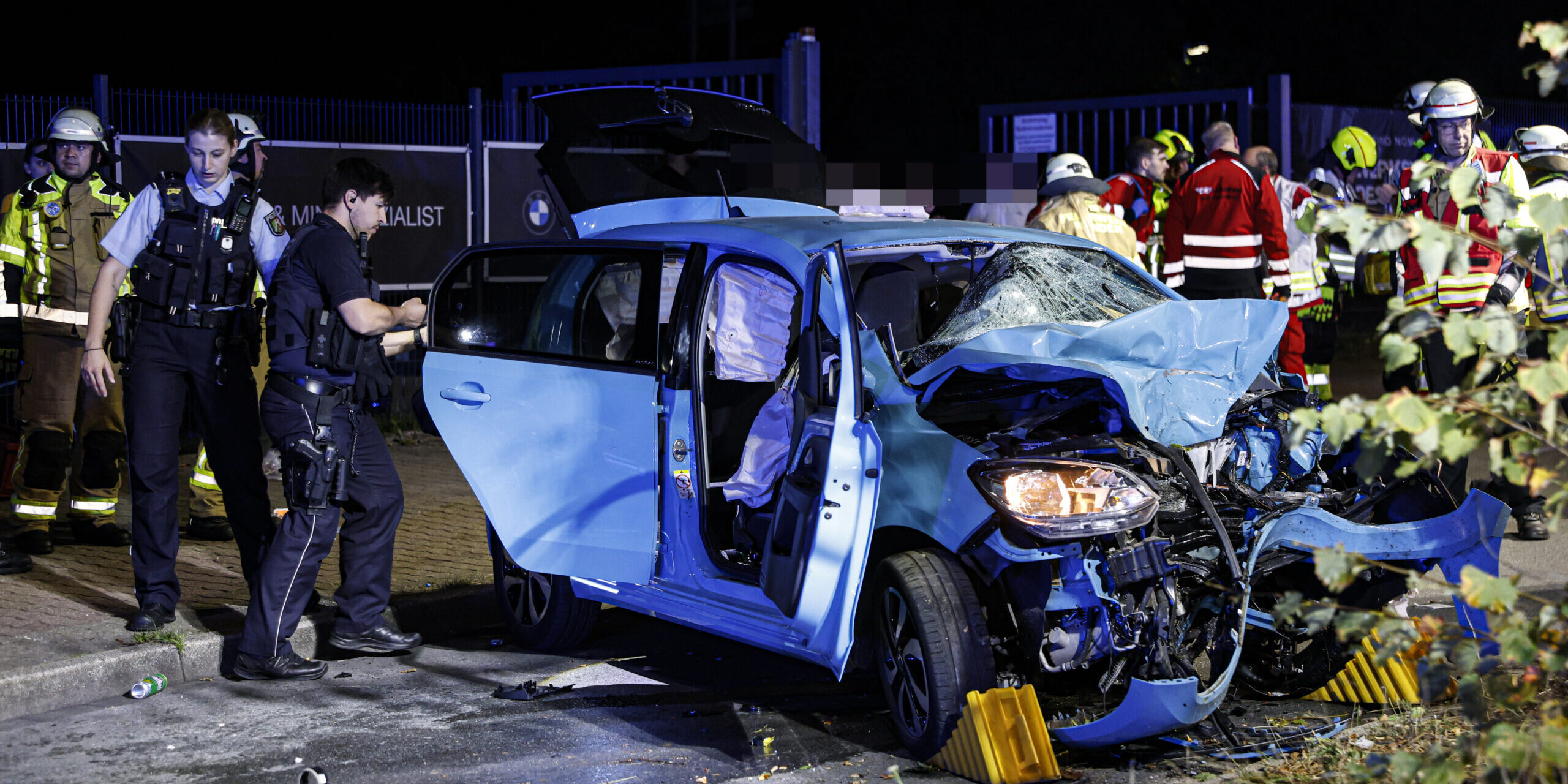 Ein blauer Kleinwagen mit völlig eingedrückter Motorpartie steht auf einer Straße, umringt von Polizisten und Feuerwehrleuten.