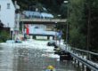 Auf einer überschwemmten Uferstraße liegen oder dümpeln mehrere Autos und Mülltonnen.