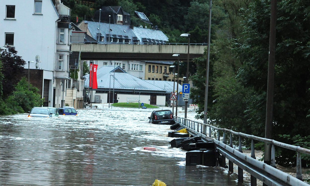 Auf einer überschwemmten Uferstraße liegen oder dümpeln mehrere Autos und Mülltonnen.