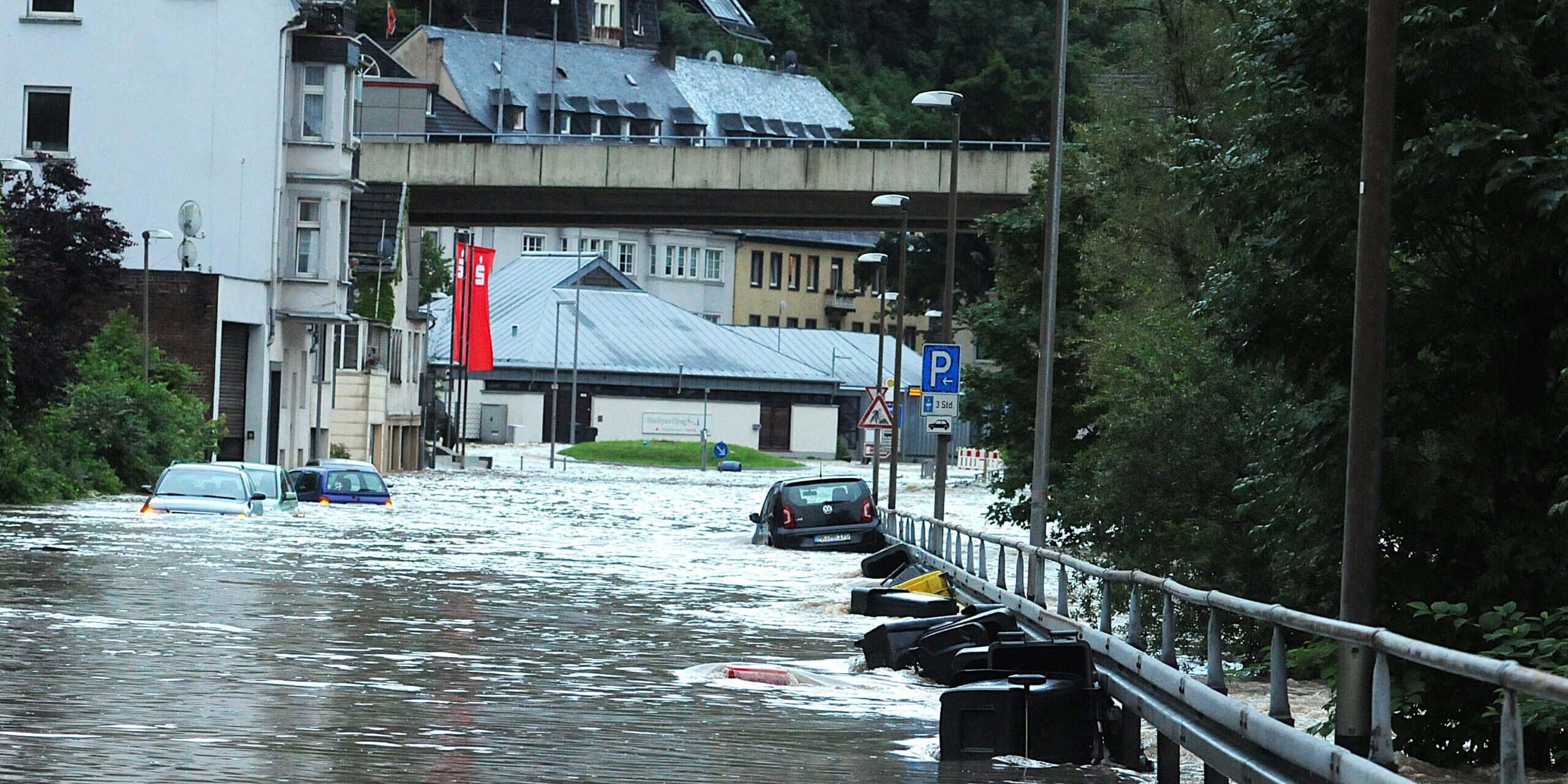 Auf einer überschwemmten Uferstraße liegen oder dümpeln mehrere Autos und Mülltonnen.
