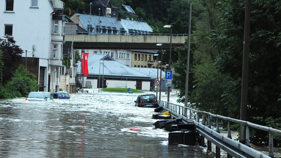 Auf einer überschwemmten Uferstraße liegen oder dümpeln mehrere Autos und Mülltonnen.