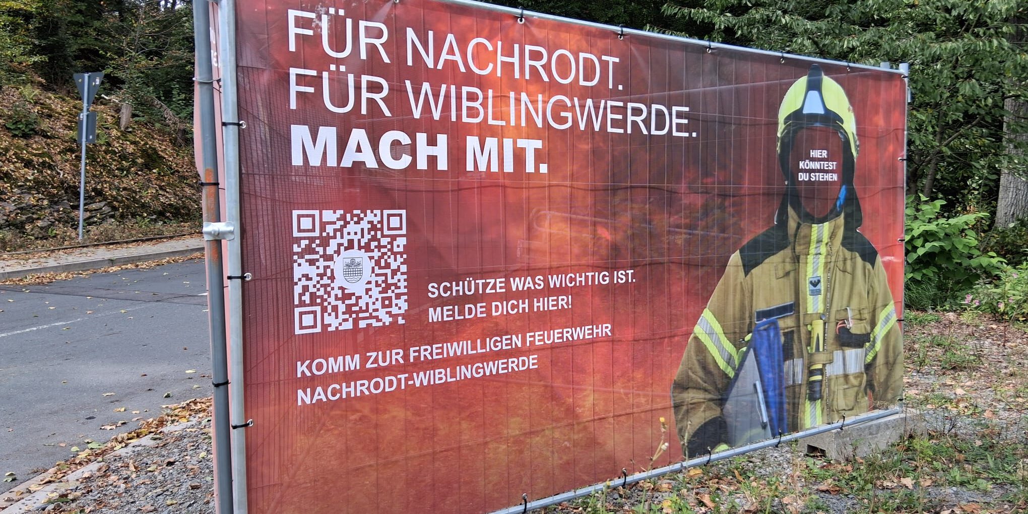 Große Werbe-Banner wie dieses am Bahnübergang Helbecke in Nachrodt machen auf die Mitglieder-Offensive der Feuerwehr aufmerksam. 
© Foto: Ilka Kremer Ein Werbeschild der Feuerwehr steht an einer Straße.