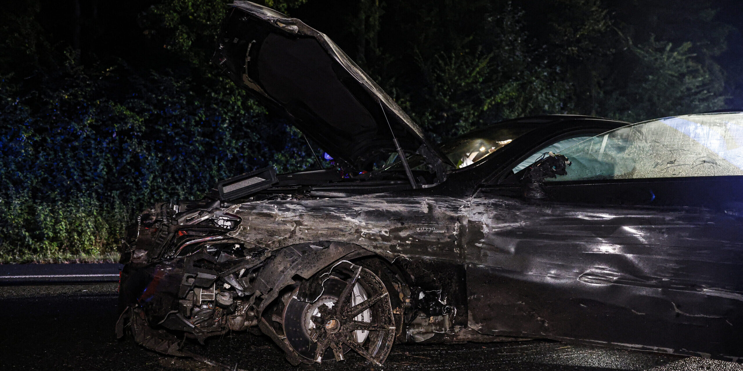 Ein dunkler Mittelklassewagen steht nachts auf einer Autobahn. Die Fahrerseite ist völlig verschrammt, die Motorhaube aufgeklappt und der linke Vorderreifen fehlt.