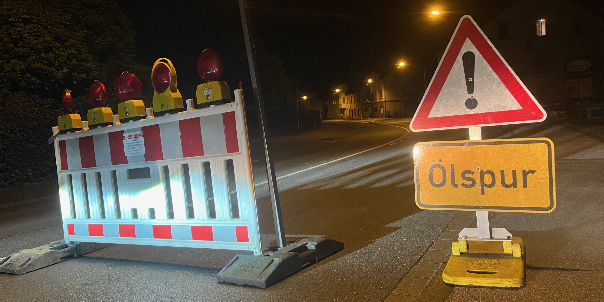 Rot-weiße Absperrbaken und ein Verkehrsschild "Achtung!" mit Zusatz "Ölspur" stehen auf einer nächtlichen Straße