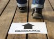 Ein Zettel mit einem Pfeil, der zur Kommunalwahl führt, liegt auf einem Holzboden.
