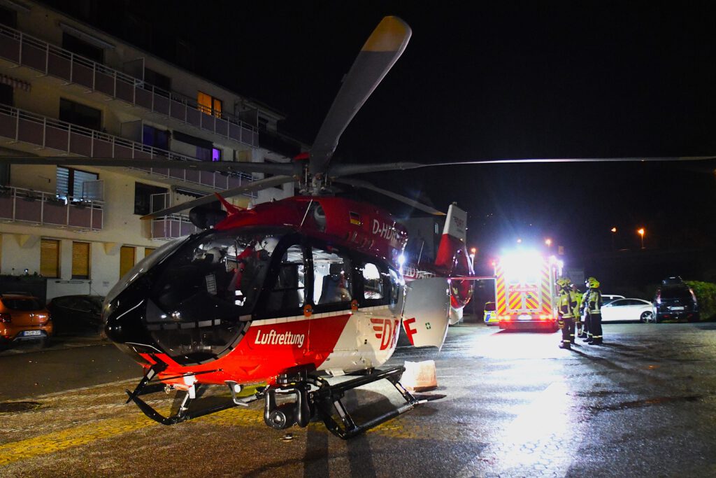 Ein rot-weißer Rettungshubschrauber steht des Nachts auf einem Landeplatz auf einem Parkplatz. Im Hintergrund stehen Feuerwehrmänner und ein Rettungswagen.