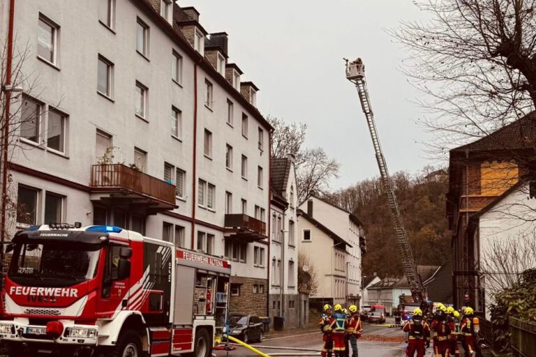 Ein Löschfahrzeug und eine ausgefahrene Drehleiter stehen auf einer Straße vor einem weiß angestrichenen Mehrfamilienhaus.