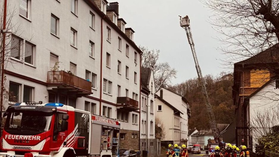 Ein Löschfahrzeug und eine ausgefahrene Drehleiter stehen auf einer Straße vor einem weiß angestrichenen Mehrfamilienhaus.