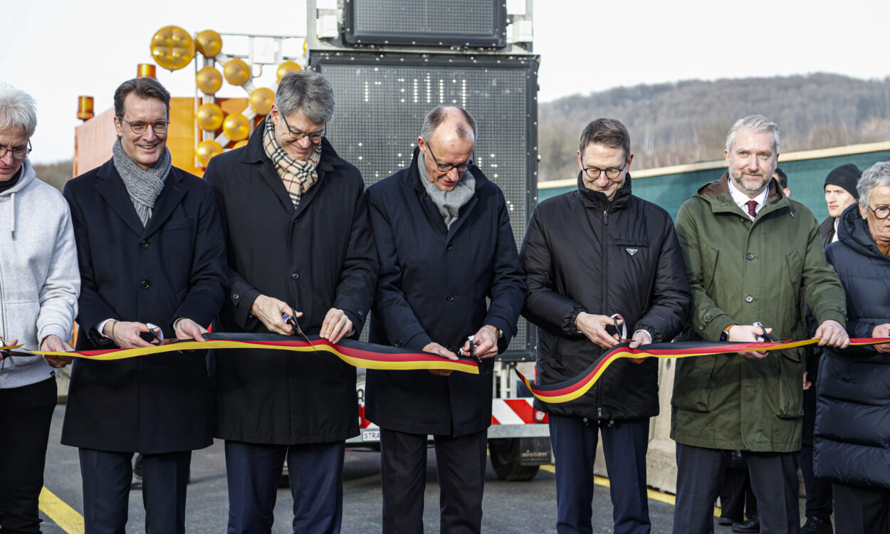 Fünf Männer, darunter Hendrik Wüst und Friedrich Merz, schneiden ein schwarz-rot-goldenes Band durch.