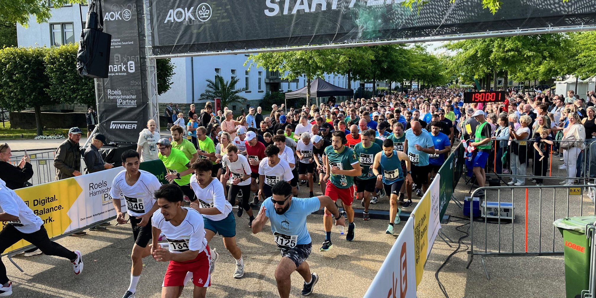 Zahlreiche Läuferinnen und Läufer starten gleichzeitig beim AOK-Firmenlauf 2025 und überqueren die Startlinie unter einem „Start/Ziel“-Banner. Zuschauer stehen dicht gedrängt hinter Absperrungen, im Hintergrund sind Bäume und die Gebäude der ehemaligen Blücher-Kaserne zu sehen.