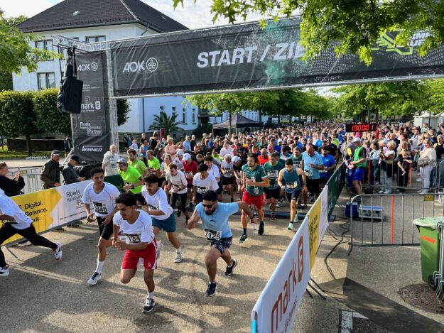 Zahlreiche Läuferinnen und Läufer starten gleichzeitig beim AOK-Firmenlauf 2025 und überqueren die Startlinie unter einem „Start/Ziel“-Banner. Zuschauer stehen dicht gedrängt hinter Absperrungen, im Hintergrund sind Bäume und die Gebäude der ehemaligen Blücher-Kaserne zu sehen.