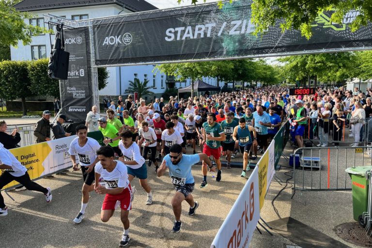 Zahlreiche Läuferinnen und Läufer starten gleichzeitig beim AOK-Firmenlauf 2025 und überqueren die Startlinie unter einem „Start/Ziel“-Banner. Zuschauer stehen dicht gedrängt hinter Absperrungen, im Hintergrund sind Bäume und die Gebäude der ehemaligen Blücher-Kaserne zu sehen.