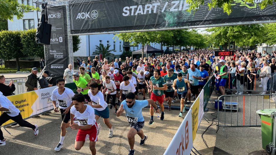 Zahlreiche Läuferinnen und Läufer starten gleichzeitig beim AOK-Firmenlauf 2025 und überqueren die Startlinie unter einem „Start/Ziel“-Banner. Zuschauer stehen dicht gedrängt hinter Absperrungen, im Hintergrund sind Bäume und die Gebäude der ehemaligen Blücher-Kaserne zu sehen.