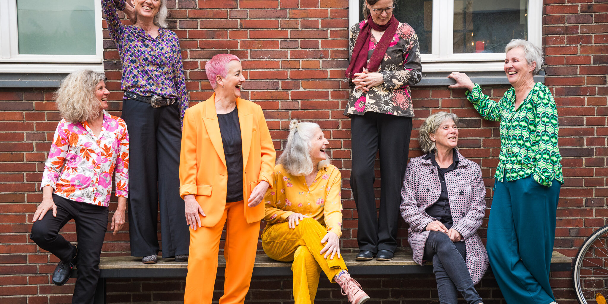 Acht ältere Frauen in bunten Outfits stehen und sitzen lachend auf einer Bank vor einer roten Backsteinwand. Sie wirken entspannt, unterhalten sich miteinander und strahlen Lebensfreude aus.