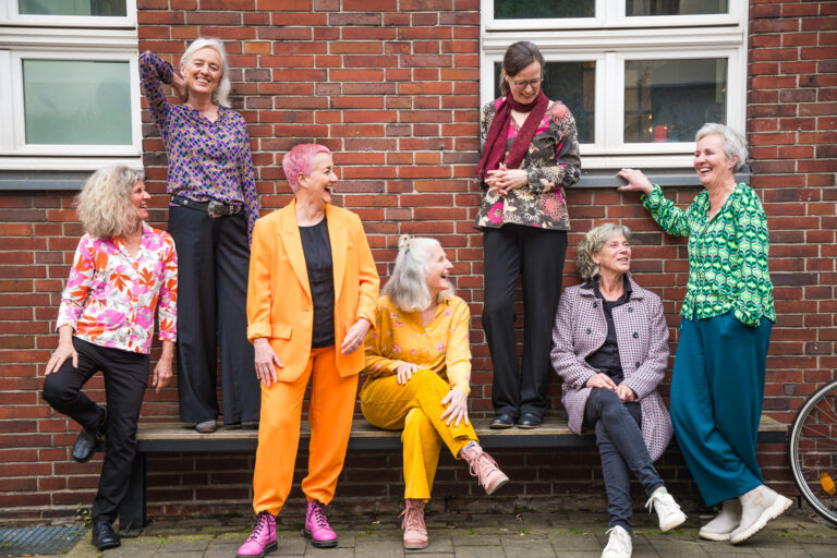 Foto: Mika Latour Acht ältere Frauen in bunten Outfits stehen und sitzen lachend auf einer Bank vor einer roten Backsteinwand. Sie wirken entspannt, unterhalten sich miteinander und strahlen Lebensfreude aus.