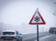 Autos auf einer verschneiten Straße, im Vordergrund ein Schild, das vor Glätte warnt