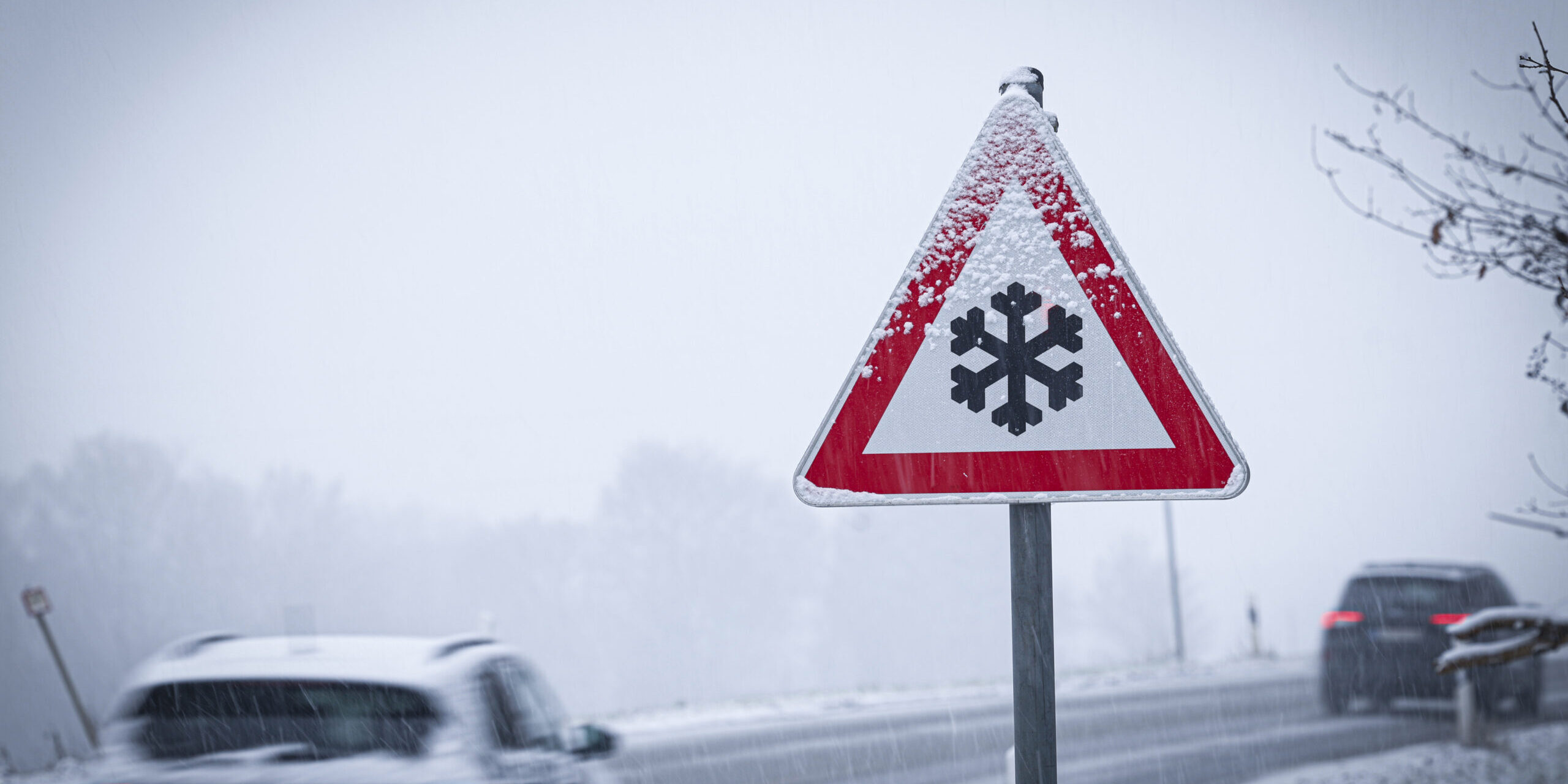 Autos auf einer verschneiten Straße, im Vordergrund ein Schild, das vor Glätte warnt