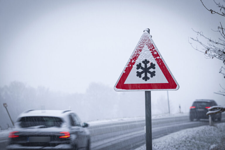 Autos auf einer verschneiten Straße, im Vordergrund ein Schild, das vor Glätte warnt