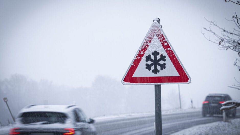 Autos auf einer verschneiten Straße, im Vordergrund ein Schild, das vor Glätte warnt