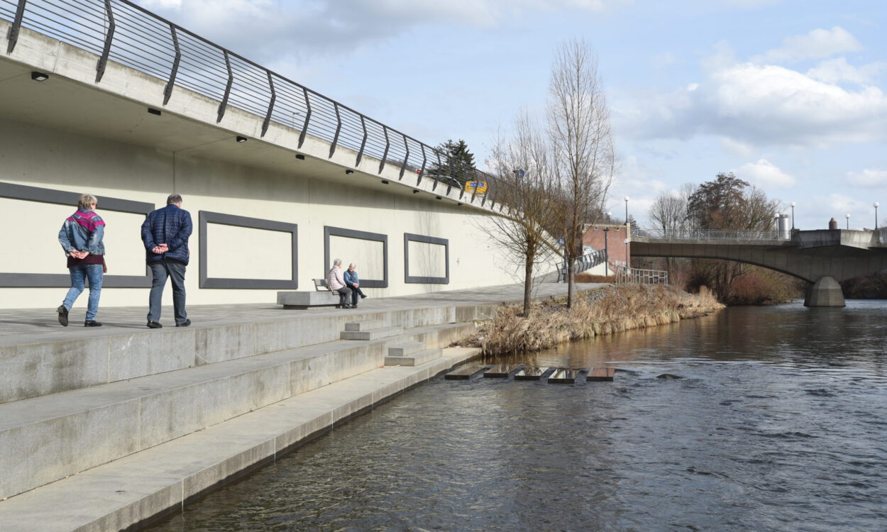Mehrere ältere Menschen spazieren und sitzen auf einer breiten Uferpromenade entlang der Lenne in Letmathe. Links verläuft eine erhöhte Straße mit Geländer, rechts fließt das Wasser, im Hintergrund sind eine Brücke, Bäume und ein teils bewölkter Himmel zu sehen.