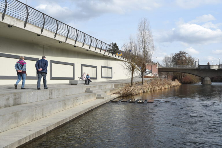 Foto: Alexander Barth Mehrere ältere Menschen spazieren und sitzen auf einer breiten Uferpromenade entlang der Lenne in Letmathe. Links verläuft eine erhöhte Straße mit Geländer, rechts fließt das Wasser, im Hintergrund sind eine Brücke, Bäume und ein teils bewölkter Himmel zu sehen.