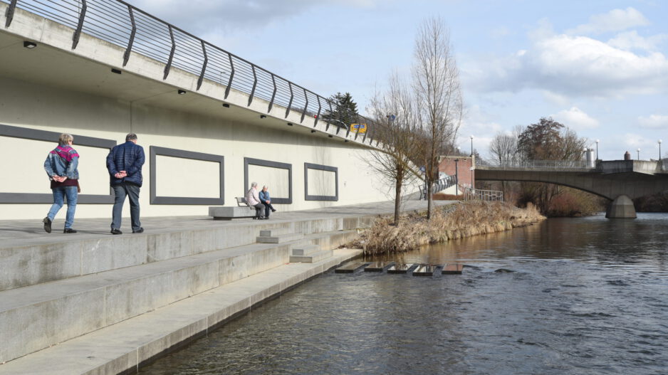 Mehrere ältere Menschen spazieren und sitzen auf einer breiten Uferpromenade entlang der Lenne in Letmathe. Links verläuft eine erhöhte Straße mit Geländer, rechts fließt das Wasser, im Hintergrund sind eine Brücke, Bäume und ein teils bewölkter Himmel zu sehen.