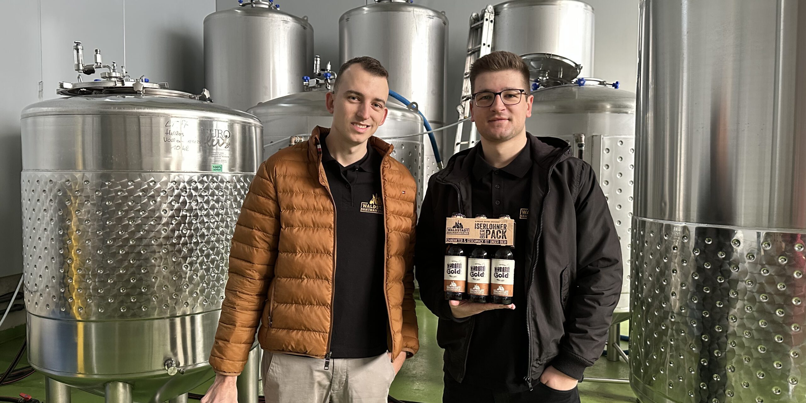 Devin Gerdes (li.) und Daniel Jürck stehen vor silbernen Tanks der Waldstadt-Braumanufaktur.