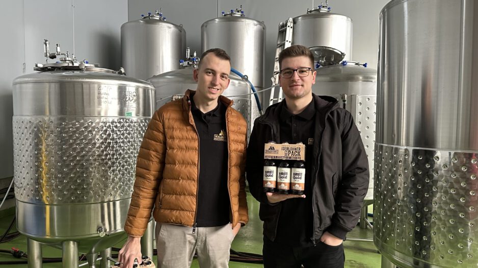 Devin Gerdes (li.) und Daniel Jürck stehen vor silbernen Tanks der Waldstadt-Braumanufaktur.