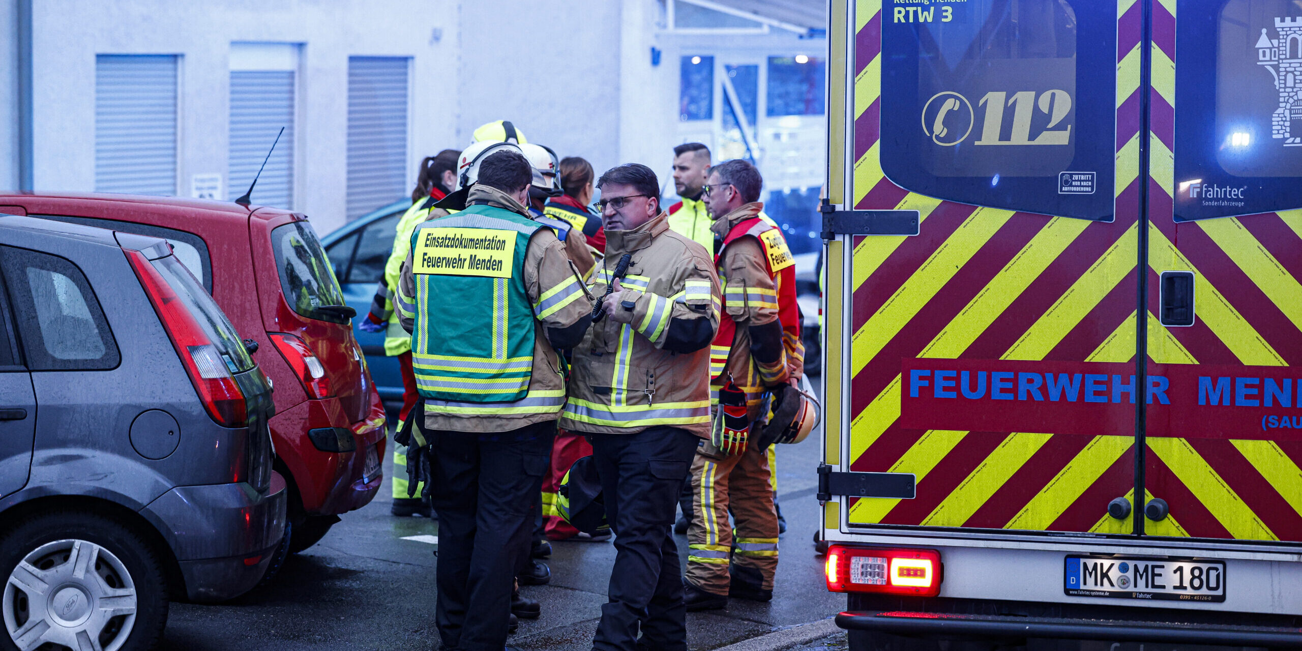 Feuerwehrleute stehen vor einem Wohnhaus, am rechten Bildrand sieht man einen Rettungswagen.