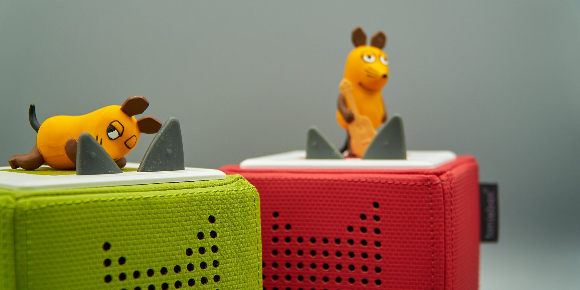 Zwei kleine Spielfiguren in Form der Maus aus der "Sendung mit der Maus" stehen auf bunten, quaderförmigen Lautsprechern (Tonieboxen). Eine Maus liegt auf einem grünen Lautsprecher, die andere steht auf einem roten, beide vor grauem Hintergrund.