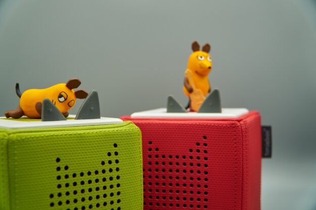 Zwei kleine Spielfiguren in Form der Maus aus der "Sendung mit der Maus" stehen auf bunten, quaderförmigen Lautsprechern (Tonieboxen). Eine Maus liegt auf einem grünen Lautsprecher, die andere steht auf einem roten, beide vor grauem Hintergrund.