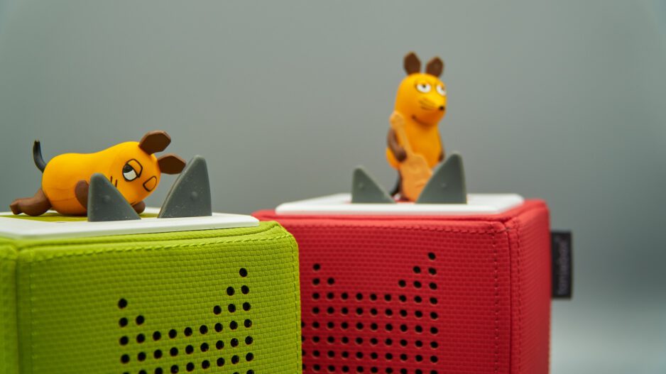 Zwei kleine Spielfiguren in Form der Maus aus der "Sendung mit der Maus" stehen auf bunten, quaderförmigen Lautsprechern (Tonieboxen). Eine Maus liegt auf einem grünen Lautsprecher, die andere steht auf einem roten, beide vor grauem Hintergrund.