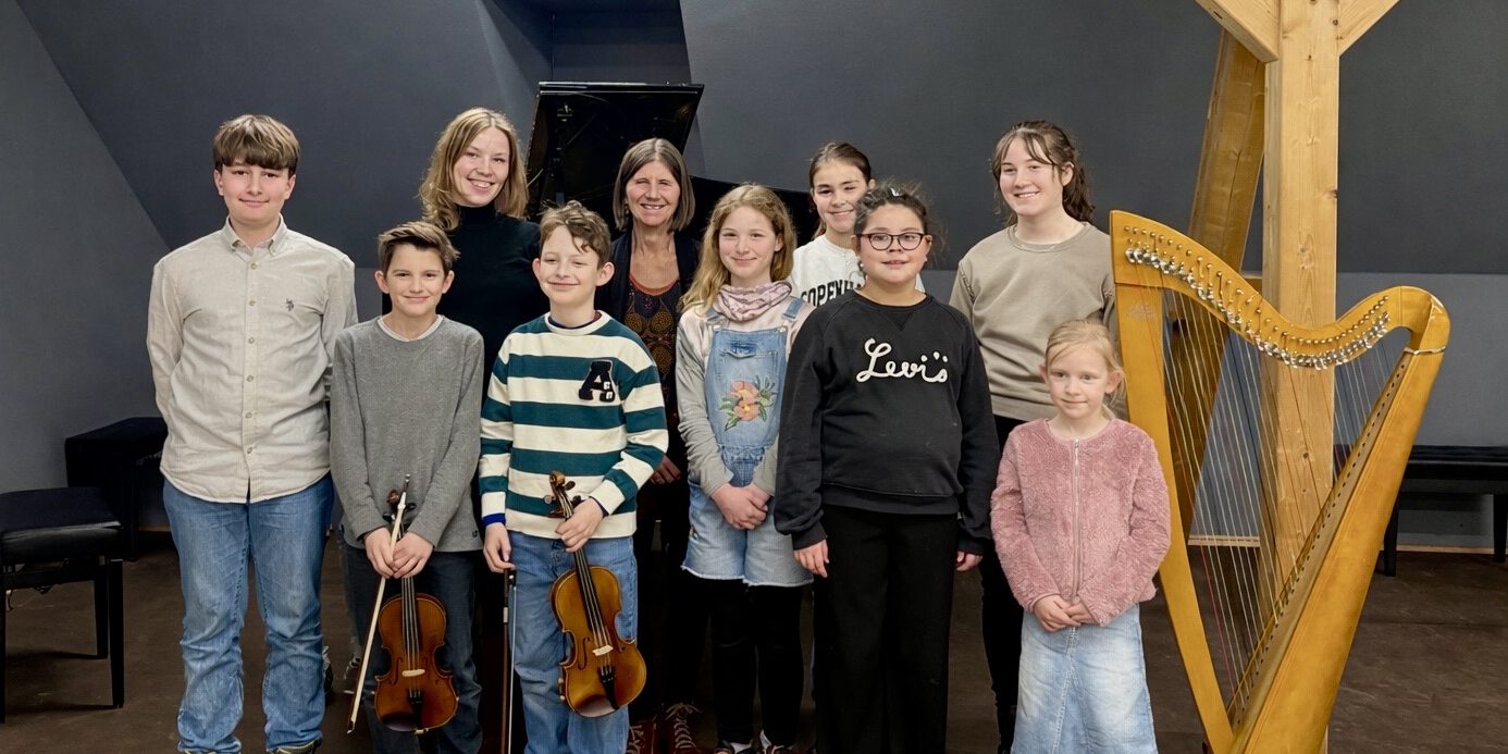 Gruppe von Kindern und zwei erwachsenen Frauen steht in einem Musikraum vor einem Klavier; einige Kinder halten Geigen, daneben steht eine Harfe.