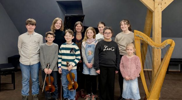 Gruppe von Kindern und zwei erwachsenen Frauen steht in einem Musikraum vor einem Klavier; einige Kinder halten Geigen, daneben steht eine Harfe.