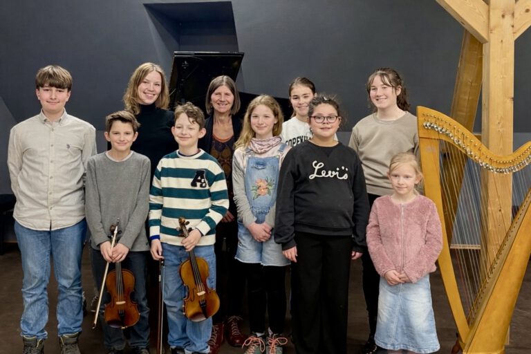Gruppe von Kindern und zwei erwachsenen Frauen steht in einem Musikraum vor einem Klavier; einige Kinder halten Geigen, daneben steht eine Harfe.