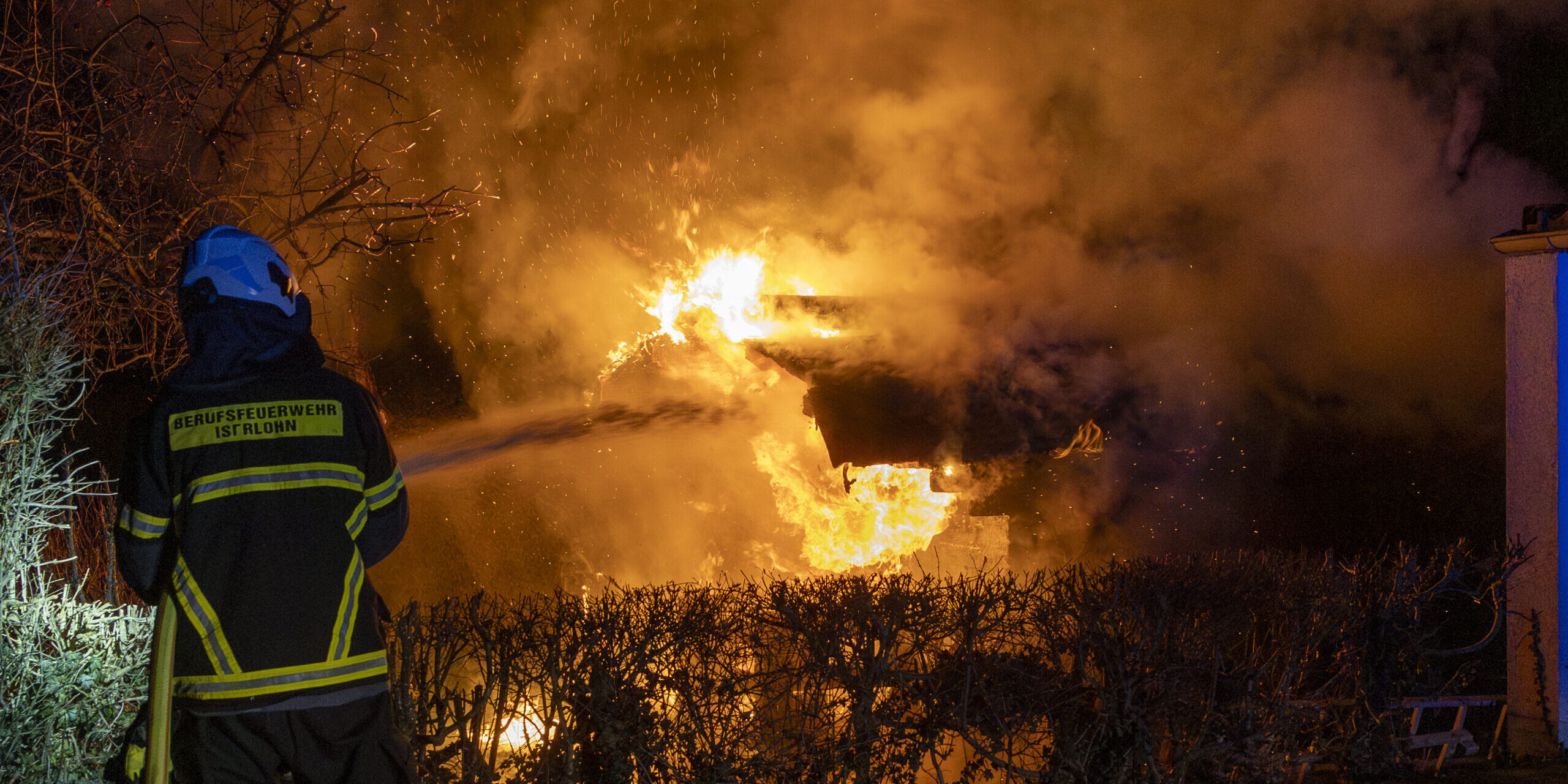 Ein Feuerwehrmann der Berufsfeuerwehr steht mit Schutzkleidung und Helm vor einem brennenden Gebäude. Hohe Flammen und dichter Rauch erleuchten die Nacht, Büsche im Vordergrund sind vom Feuer umgeben.