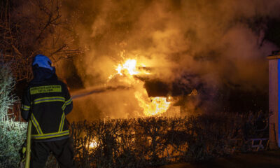 Ein Feuerwehrmann der Berufsfeuerwehr steht mit Schutzkleidung und Helm vor einem brennenden Gebäude. Hohe Flammen und dichter Rauch erleuchten die Nacht, Büsche im Vordergrund sind vom Feuer umgeben.