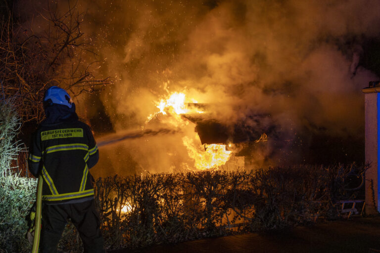 Ein Feuerwehrmann der Berufsfeuerwehr steht mit Schutzkleidung und Helm vor einem brennenden Gebäude. Hohe Flammen und dichter Rauch erleuchten die Nacht, Büsche im Vordergrund sind vom Feuer umgeben.