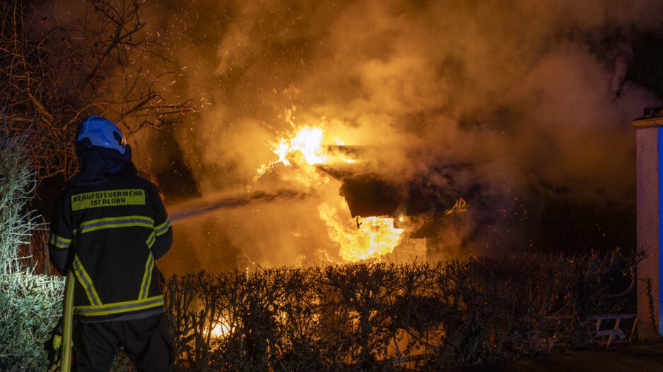 Ein Feuerwehrmann der Berufsfeuerwehr steht mit Schutzkleidung und Helm vor einem brennenden Gebäude. Hohe Flammen und dichter Rauch erleuchten die Nacht, Büsche im Vordergrund sind vom Feuer umgeben.