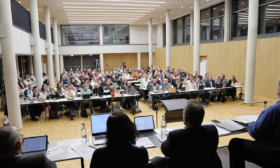 Großer Sitzungssaal mit vielen Teilnehmenden an langen Tischen; zahlreiche Personen heben die Hand während einer Abstimmung, vorne sitzt das Präsidium mit Laptops und Unterlagen.