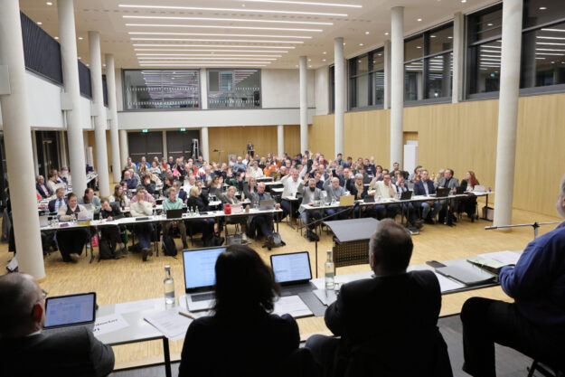 Foto: Stephan Greitzke Großer Sitzungssaal mit vielen Teilnehmenden an langen Tischen; zahlreiche Personen heben die Hand während einer Abstimmung, vorne sitzt das Präsidium mit Laptops und Unterlagen.