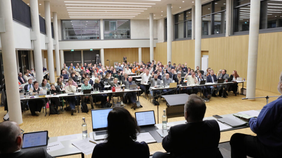 Großer Sitzungssaal mit vielen Teilnehmenden an langen Tischen; zahlreiche Personen heben die Hand während einer Abstimmung, vorne sitzt das Präsidium mit Laptops und Unterlagen.